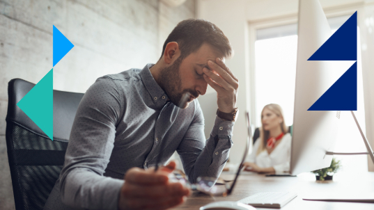 A man holds his head in frustration in front of his computer, behind him sits a woman working on a desk.